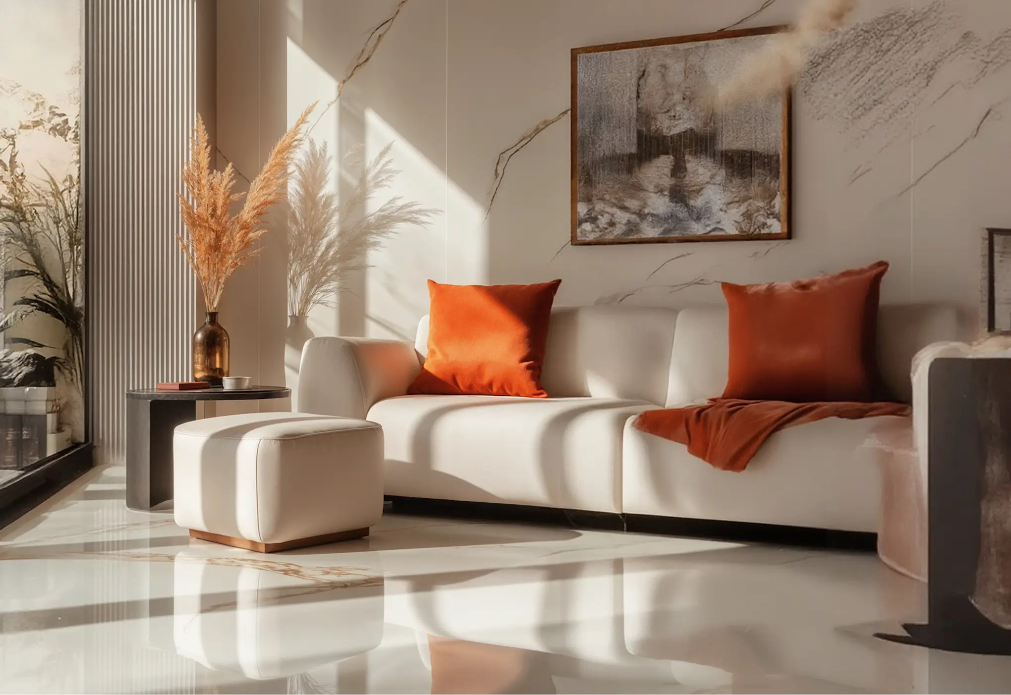 Modern living room corner with a white sectional sofa featuring two burnt orange cushions and a matching throw, a white ottoman, a small black side table with a brown vase holding dried pampas grass, and sunlight casting soft shadows on glossy marble flooring and beige walls with subtle marble veining and framed artwork.