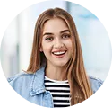 Smiling young woman with long straight light brown hair wearing a denim jacket and striped shirt, indoors with a blurred bright background.