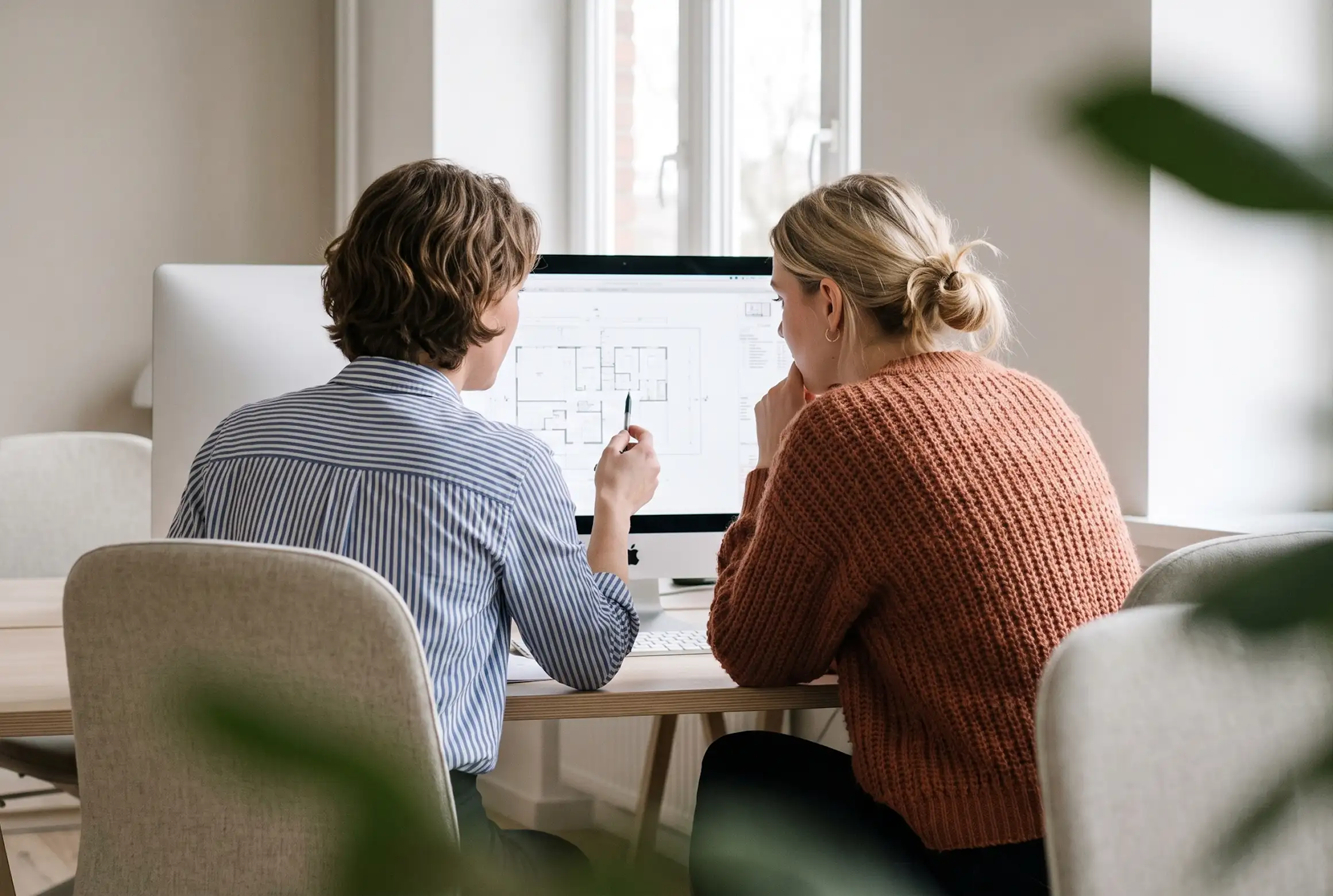 Two people sitting at a desk looking at a desktop computer screen displaying a floor plan; one person points at the screen with a pen.
