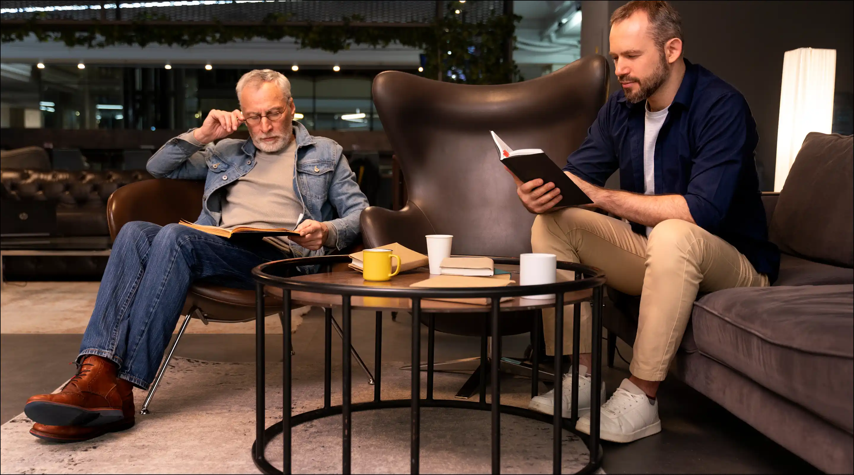 Two men sitting in a modern living room reading books; an older man in a denim jacket and jeans on a brown chair, and a younger man in a dark shirt and khaki pants on a gray sofa, with a round black metal and wooden coffee table holding books and mugs between them.